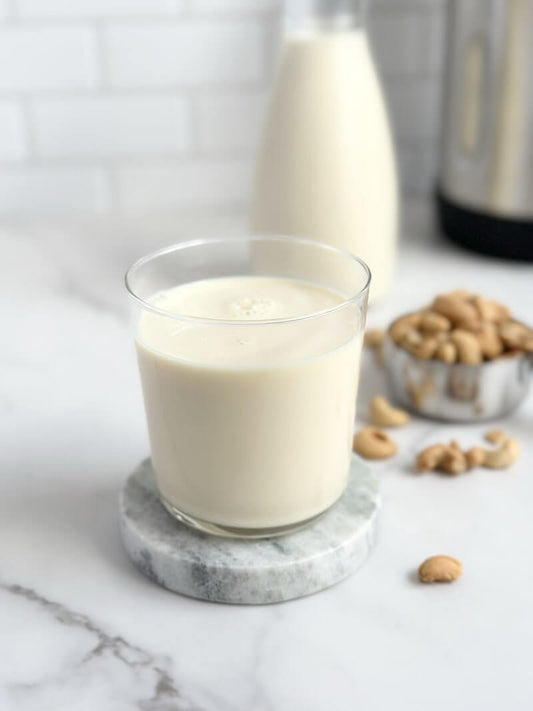 Glass of cashew milk with a bowl of raw cashew nuts in the background on a light-colored surface