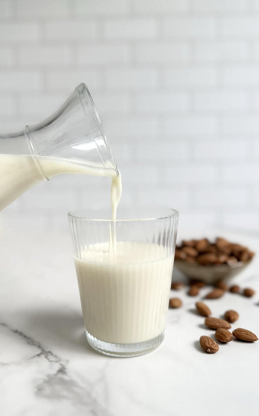 Pouring almond milk from a glass bottle into a clear glass with almonds in the background